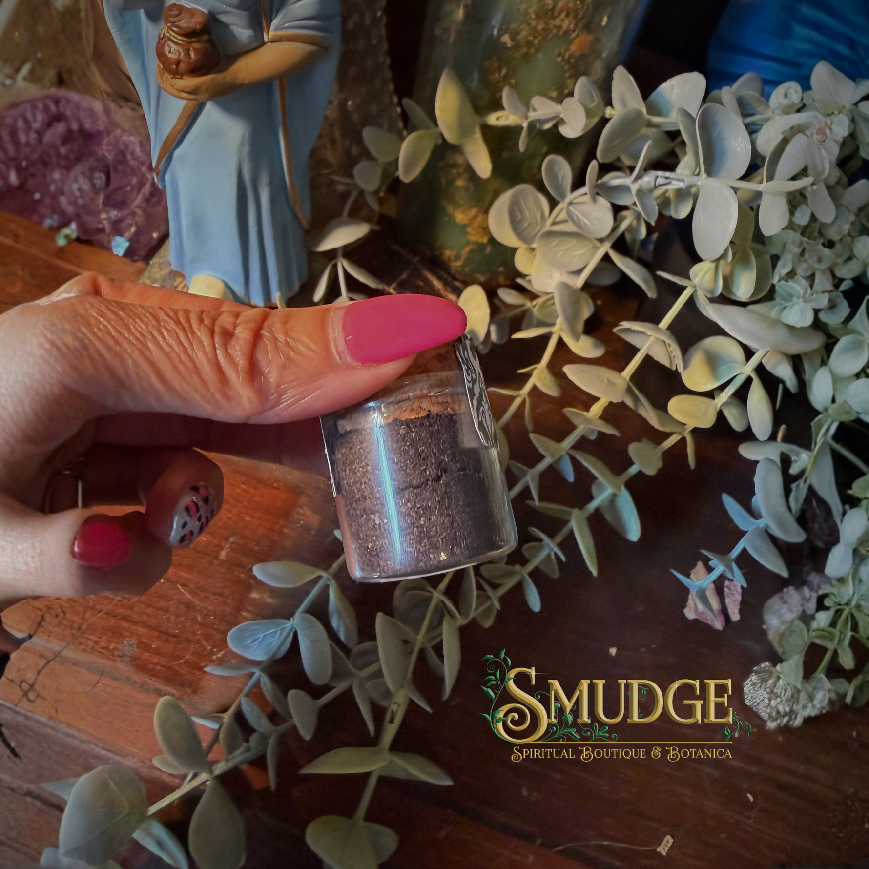 Hand holding a small glass container with herbs, surrounded by eucalyptus leaves and a figurine, on a wooden surface with 'Smudge' branding.