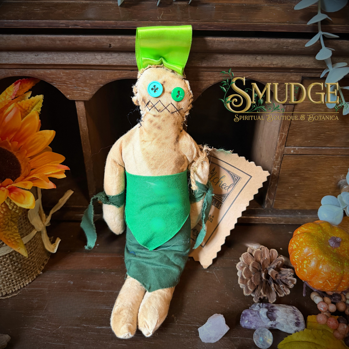 A handmade cloth doll with a beige linen body, green fabric apron and hat, and button eyes, displayed on a rustic wooden surface with autumn decor and crystals. The brand name 'Smudge Spiritual Boutique & Herbs' is visible on the right.