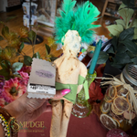 A handmade rag doll with a bright green feather headdress, green sash, and a price tag attached, held by a hand in a shop setting with flowers and decorative jars in the background.