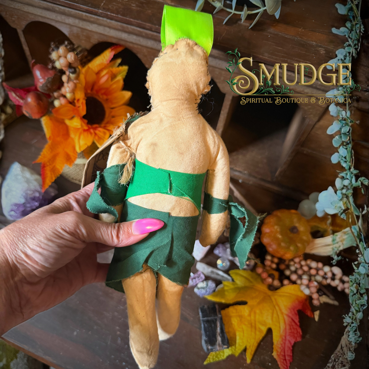 A handmade spiritual doll with a tan body, green waistcloth, and green hat is held by a hand with pink nails against a rustic autumn-themed background. The text 'SMUDGE Spiritual Boutique & Emporium' is visible in the upper right.