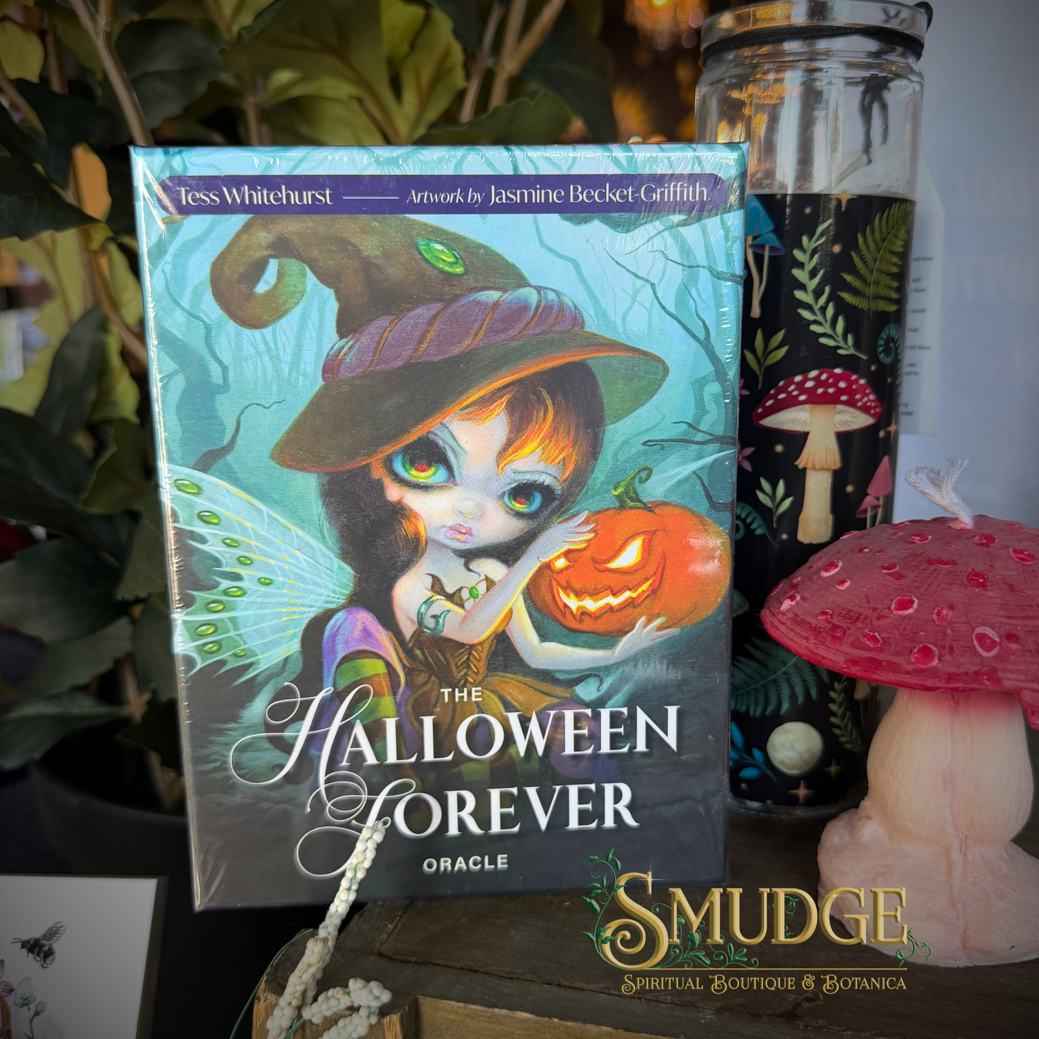 The Halloween Forever Oracle card deck box by Tess Whitehurst, illustrated by Jasmine Becket Griffith, displayed on a wooden surface next to a glass jar candle and a pink mushroom figurine. The box features a cute cartoon witch holding a jack-o'-lantern.