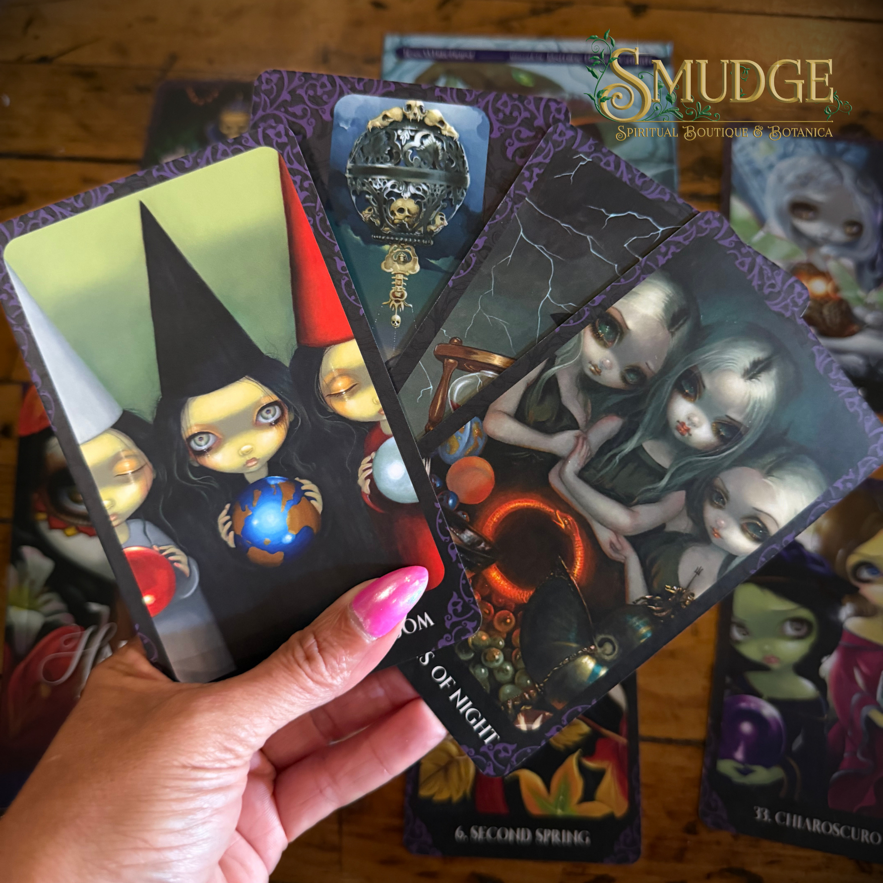 A hand with pink nail polish holds three illustrated tarot-style cards over a wooden surface, with more cards fanned out behind them. The cards feature dark fantasy artwork with pale-faced figures, purple borders, and mystical symbols.
