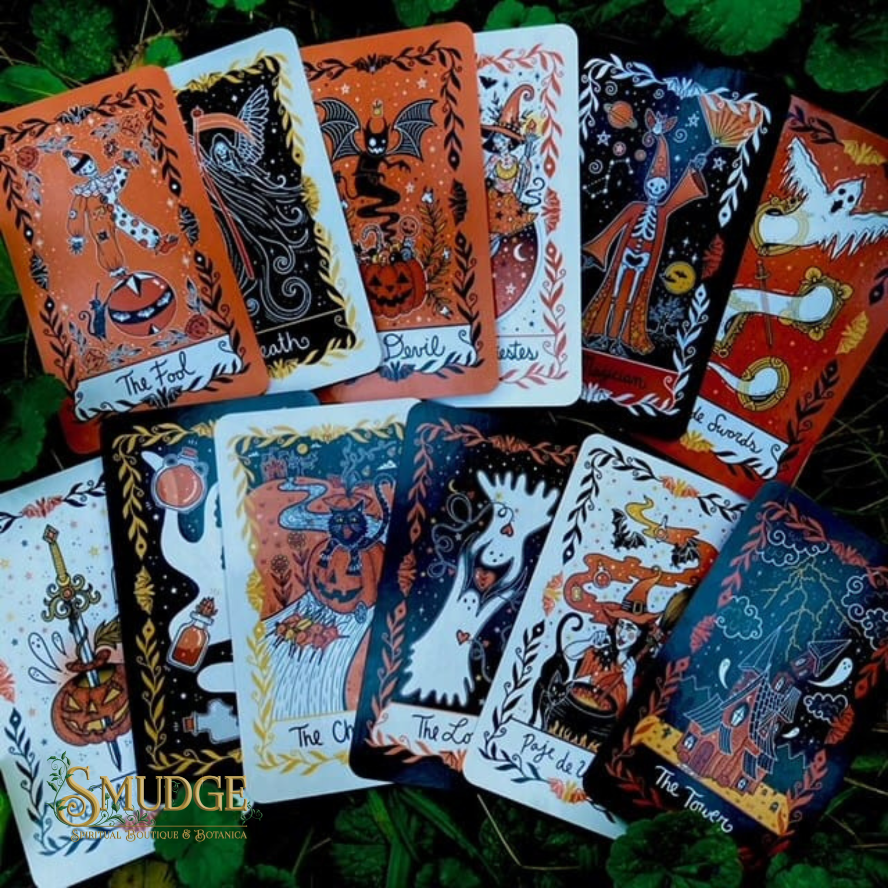 A set of illustrated tarot-style cards arranged on green foliage. The cards feature Halloween-themed artwork with pumpkins, ghosts, bats, and spooky figures in orange, black, white, and dark blue tones.