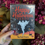 A hand holds a shrink-wrapped box of 'Happy Halloween Tarot' by Soni Graves, set against a background of pink and purple flowers. The box features a dark night sky design with a white ghost, a haunted house, and an orange ground.