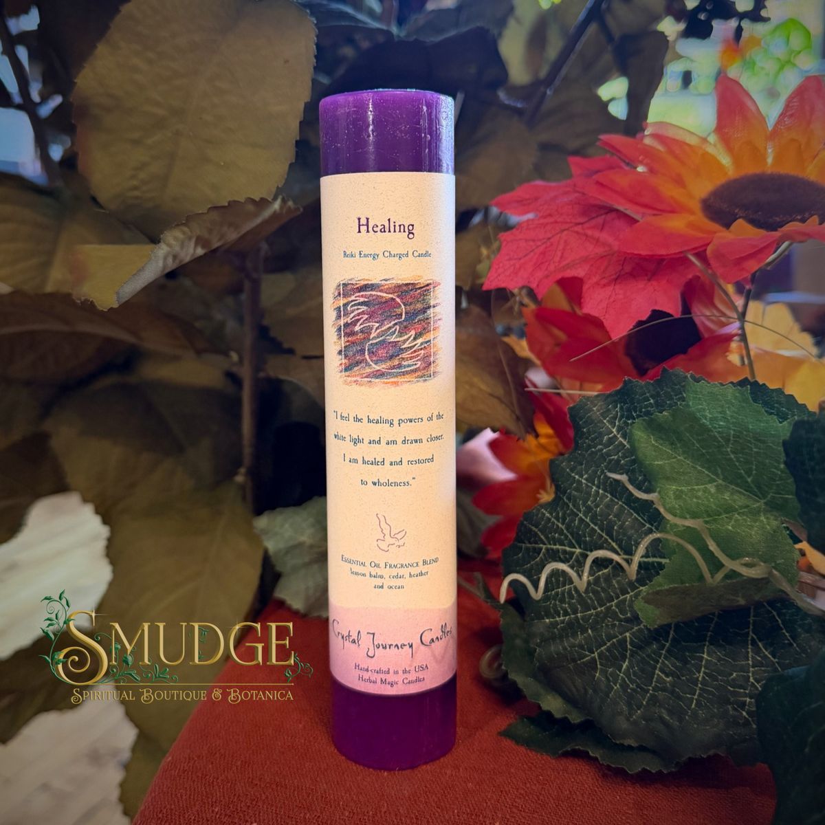 A Healing scented candle in a purple glass jar with a cream-colored label, styled with autumn leaves and flowers on a rust-colored surface. The brand 'Mudge Spiritual Boutique & Botanica' is visible as a watermark in the lower-left corner.