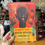 A hand with green nail polish holds a book titled 'The Healing Power of African American Spirituality' by Stephanie Rose Bird. The book cover features a colorful illustration of a Black person with a bird on a red and orange background.