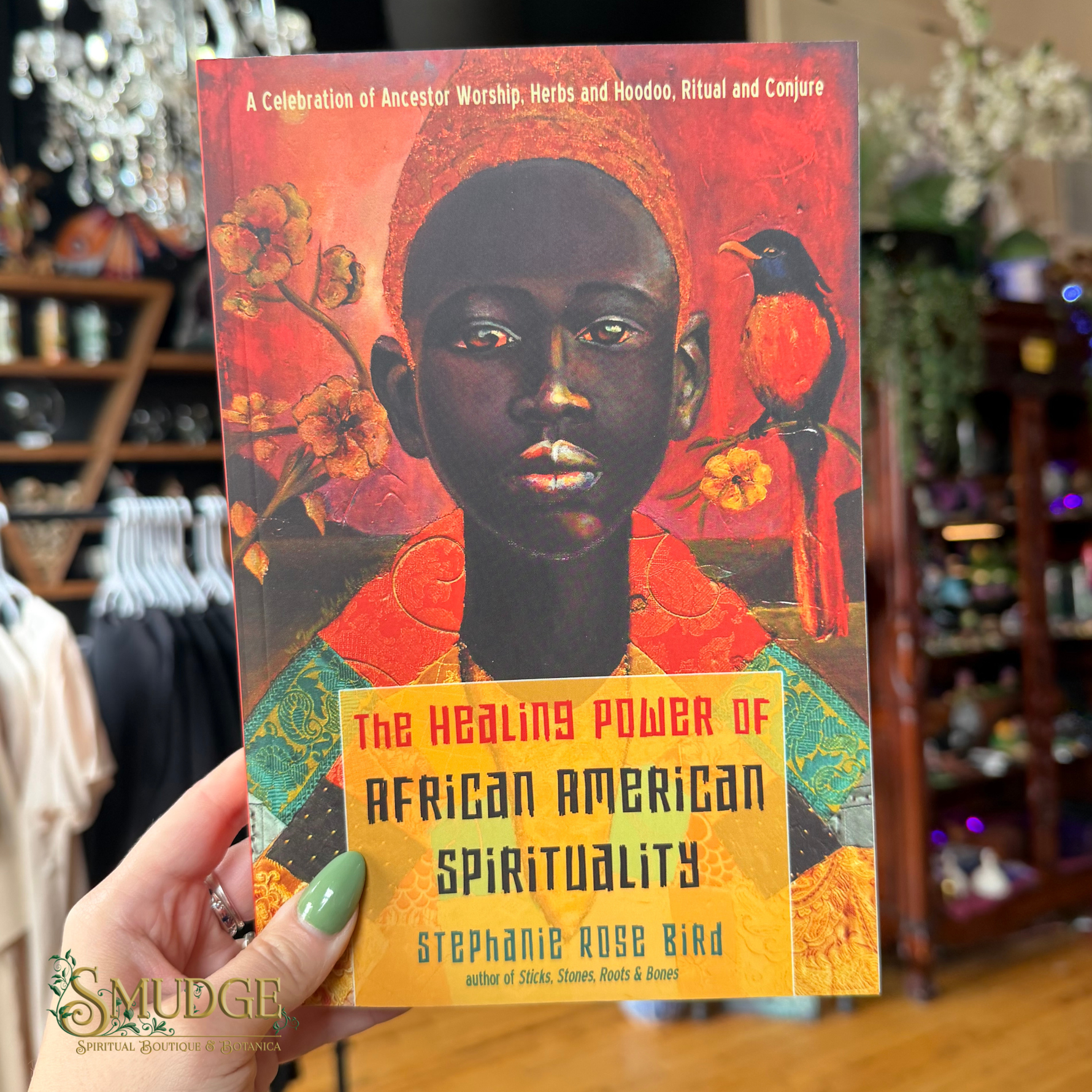 A hand with green nail polish holds a book titled 'The Healing Power of African American Spirituality' by Stephanie Rose Bird. The book cover features a colorful illustration of a Black person with a bird on a red and orange background.
