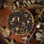 A top-down view of a brown ceramic bowl filled with loose-leaf herbal tea, surrounded by dried flowers, green leaves, and a silver spoon on a dark wooden surface. The brand logo 'The Squeeze Botanicals' is visible in the top right corner.