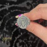 Silver ring with intricate design held by a hand against a dark textured background, featuring the brand 'Smudge'.
