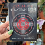 A hand with green nail polish holds a boxed card deck titled 'Oracle of the Hekatean Path' by Kenn Payne and Chris Butler. The box is dark gray/black with a red circular symbol and white and red text.
