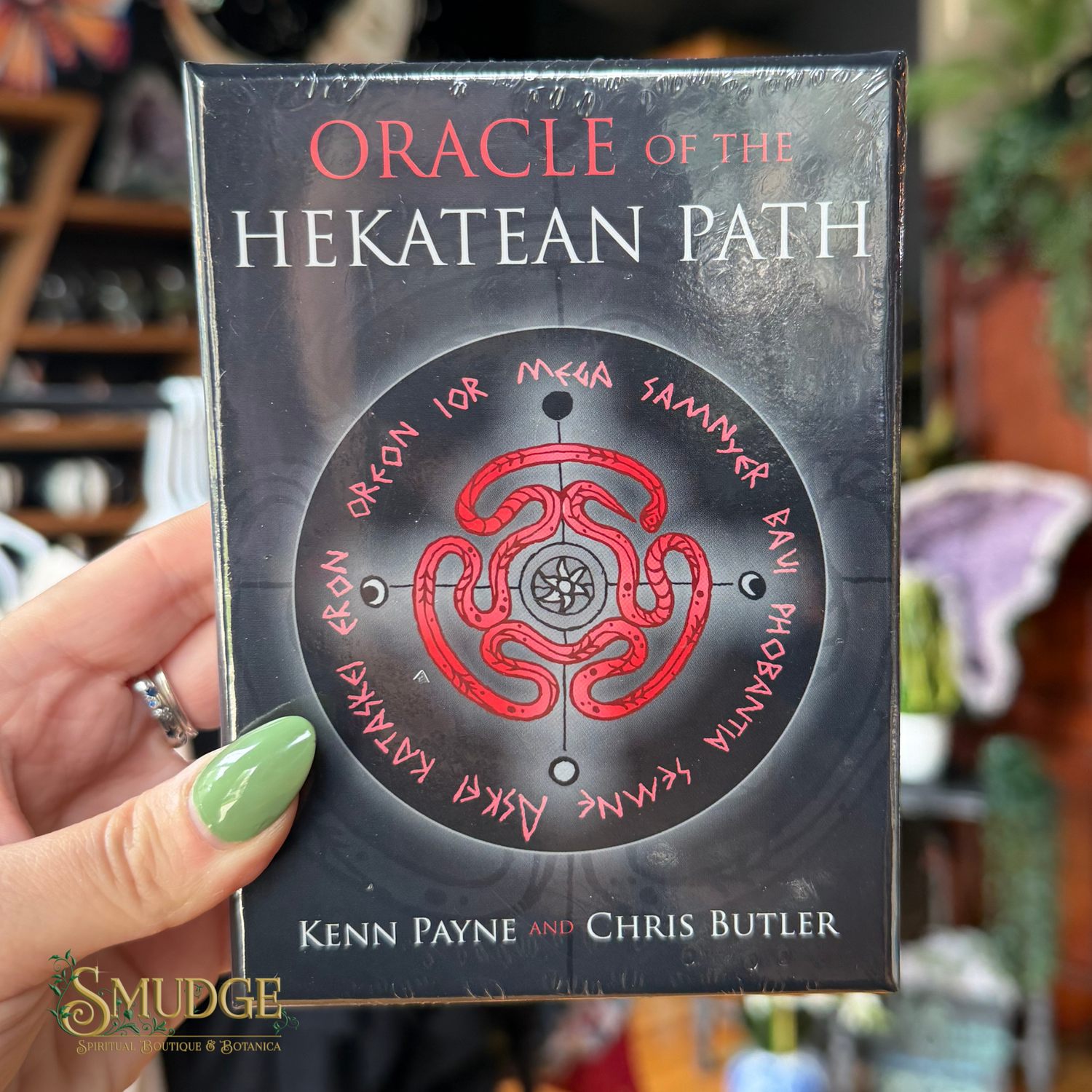 A hand with green nail polish holds a boxed card deck titled 'Oracle of the Hekatean Path' by Kenn Payne and Chris Butler. The box is dark gray/black with a red circular symbol and white and red text.