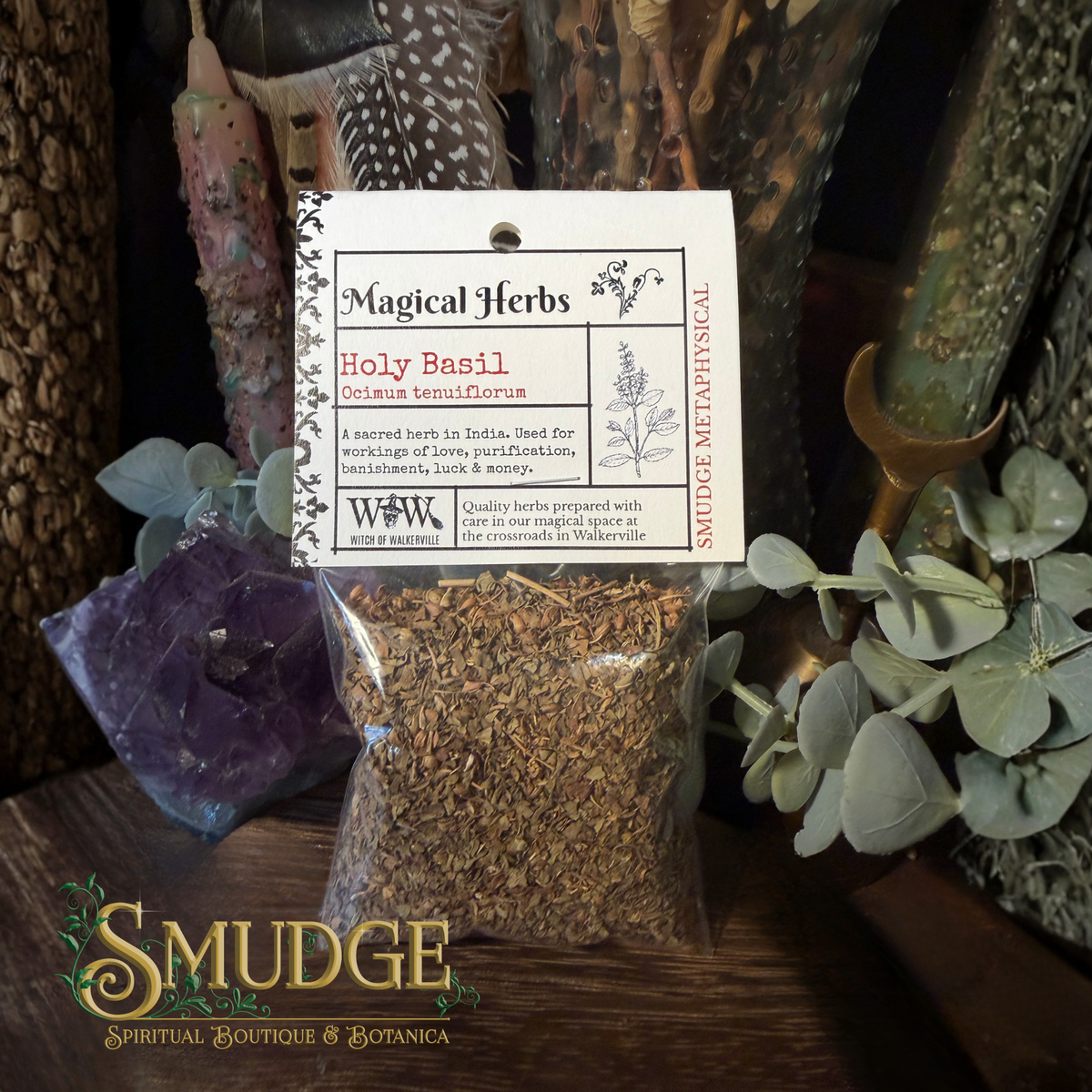 A clear plastic bag filled with dried brown herbal material sits on a wooden surface, with a white hanging label reading 'Magical Herbs' and 'Holy Basil.' The scene includes decorative plants, crystals, and a branded 'Smudge Spiritual Boutique & Botanica' logo at the bottom left.