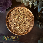 A top-down view of a wooden bowl filled with dried, shredded brown herbal or spiritual material, set on a dark wooden surface with decorative crystals, eucalyptus leaves, and metal diffusers. The brand logo 'SMUDGE Spiritual Boutique & Botanica' is visible in the bottom-left corner.