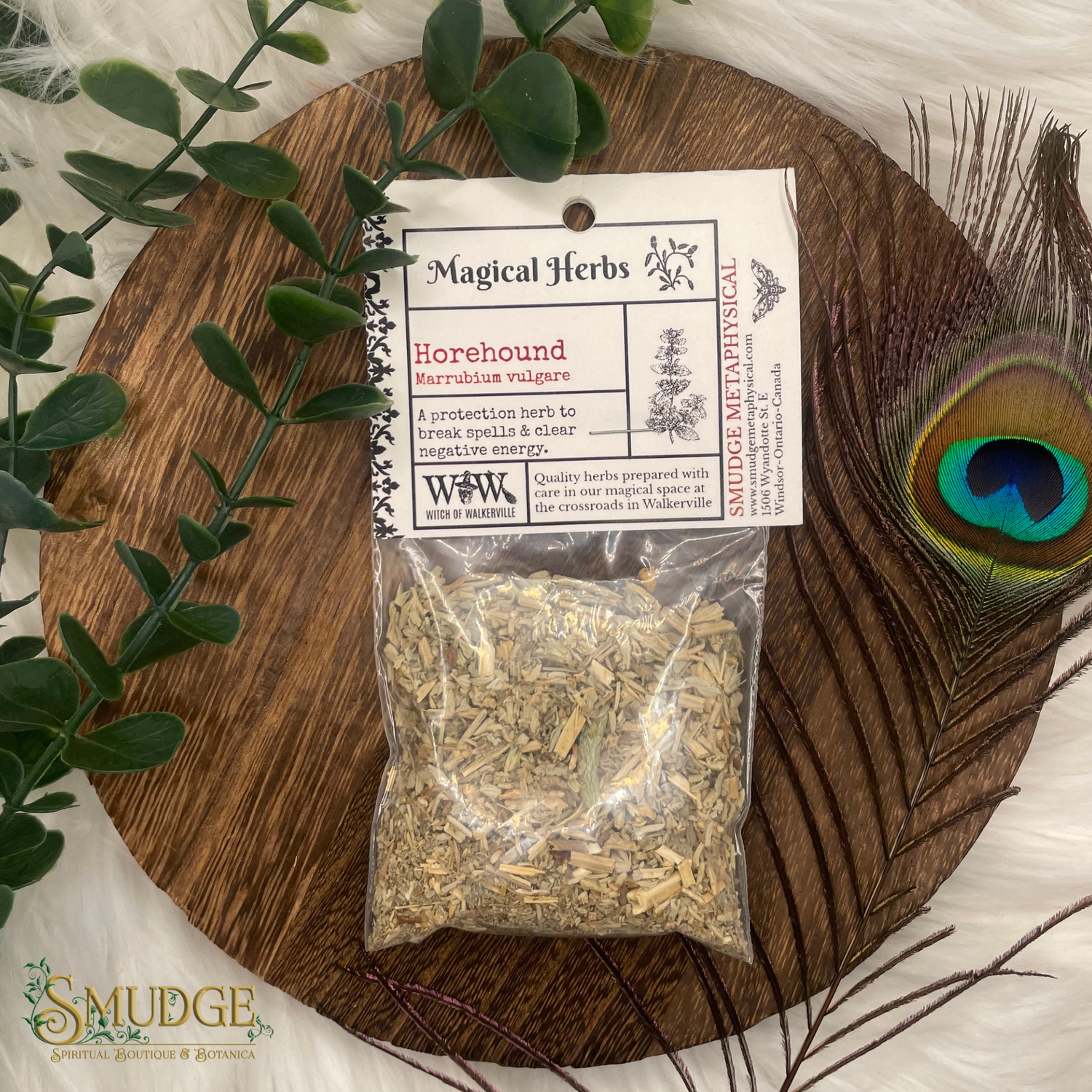 Bag of horehound herbs with a label on a wooden board, surrounded by greenery and a peacock feather.