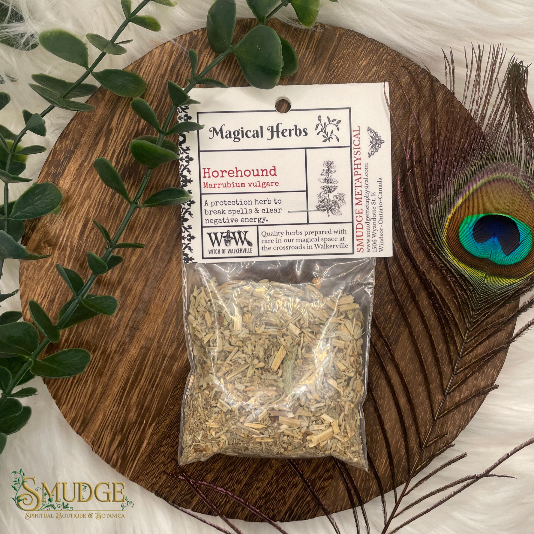 Bag of horehound herbs with a label on a wooden board, surrounded by greenery and a peacock feather.