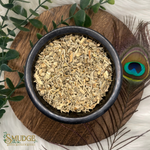 Dried herbs in a black bowl on a wooden surface with greenery and a peacock feather.