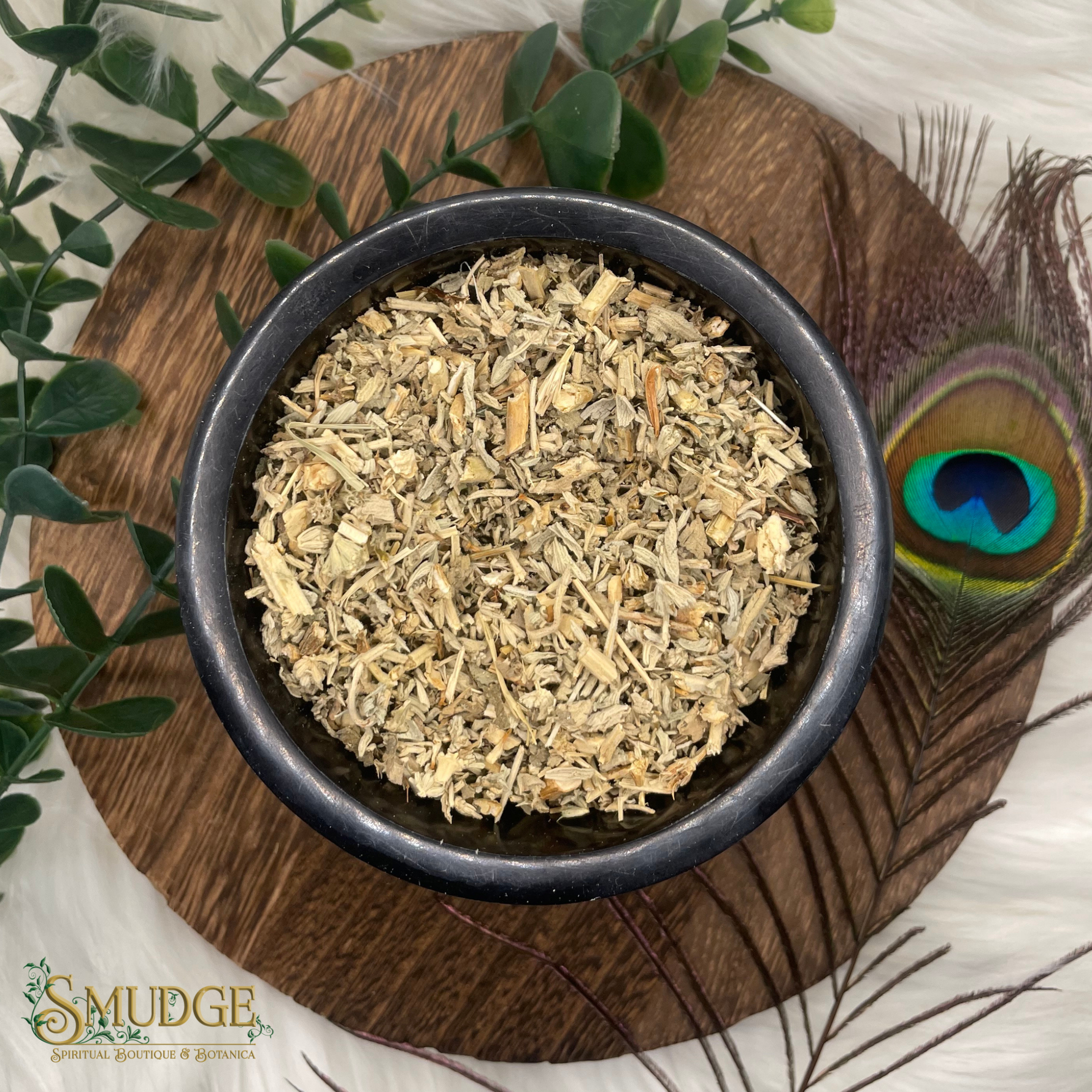 Dried herbs in a black bowl on a wooden surface with greenery and a peacock feather.