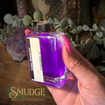 A hand with pink nail polish holds a small square glass bottle filled with bright purple liquid. The bottle has a black cap, a white label reading 'Smudler Anomalous', and is photographed over a wooden surface with plants, crystals, and a decorative 'Smudge Spiritual Boutique & Botanica' watermark at the bottom.
