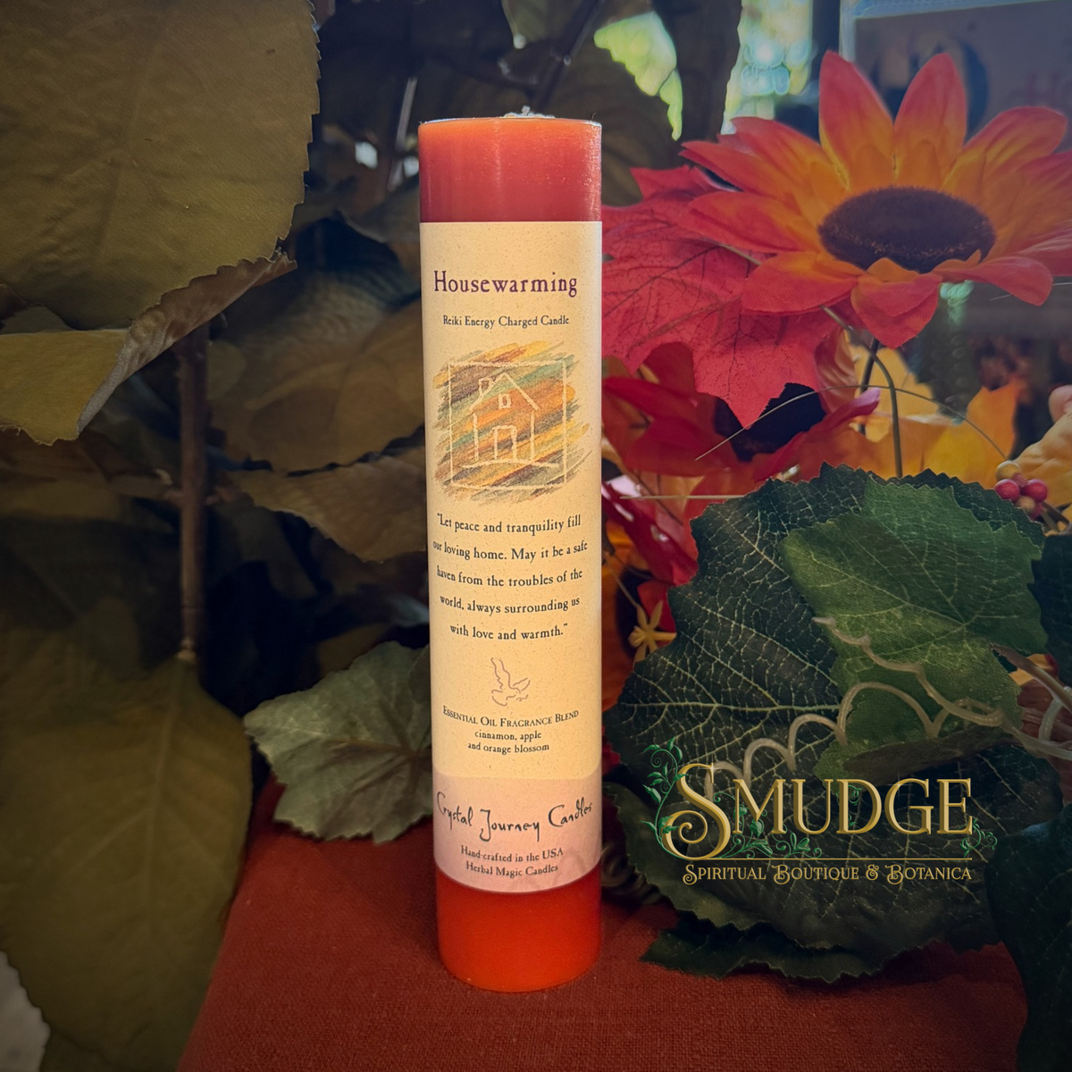 A single orange taper candle with a cream label reading 'Housewarming' and 'Sib Energy Charged Candles,' styled with autumn leaves, a sunflower, and a red cloth background. A 'SMUDGE Spiritual Boutique & Botanica' watermark appears on the lower right.
