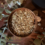 A top-down view of a rustic brown wooden bowl filled with dried shredded herbal or paper-like material, placed on a dark wooden surface with decorative leaves and stones around it. The brand name 'SMUDGE' is visible as an overlay at the bottom left.