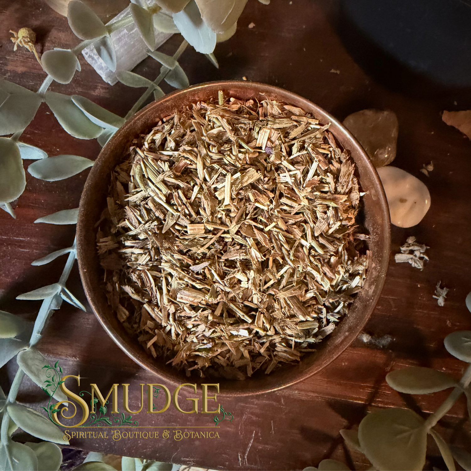 A top-down view of a rustic brown wooden bowl filled with dried shredded herbal or paper-like material, placed on a dark wooden surface with decorative leaves and stones around it. The brand name 'SMUDGE' is visible as an overlay at the bottom left.