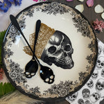 Two black spoons with skull-shaped handles resting on a decorative white plate with black floral patterns, a gold bandana print, and a black-and-white skull illustration.