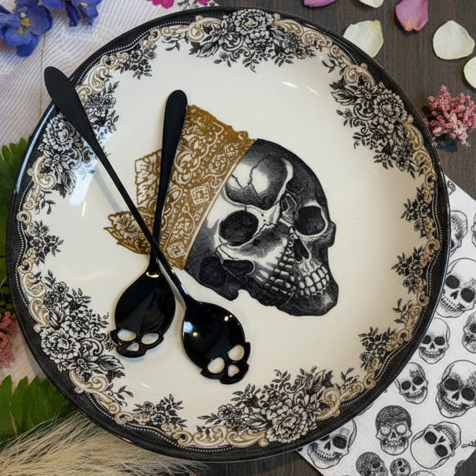 Two black spoons with skull-shaped handles resting on a decorative white plate with black floral patterns, a gold bandana print, and a black-and-white skull illustration.