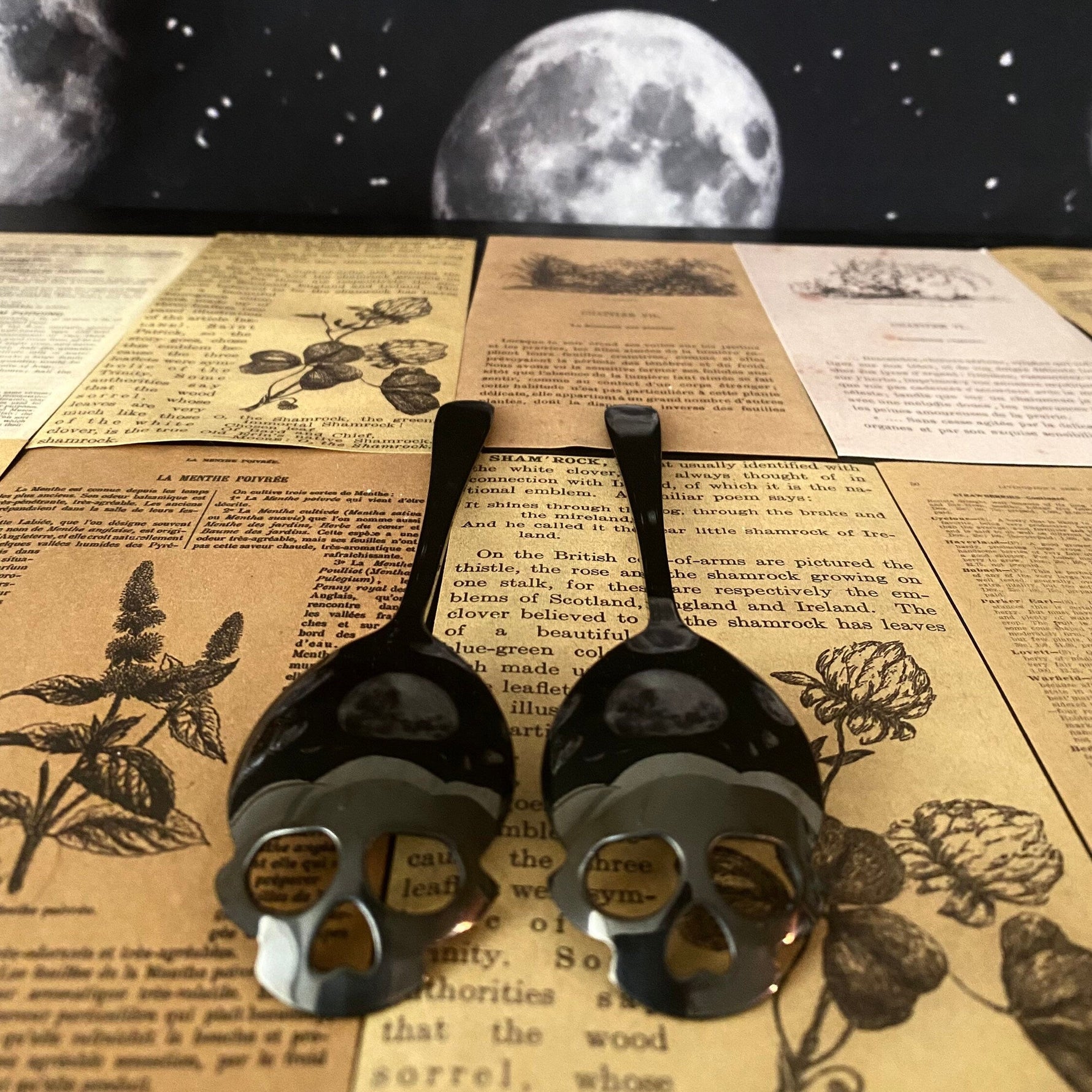 Two black metal skull-shaped spoons resting on vintage-style printed book pages, with a dark starry sky backdrop featuring moon illustrations.
