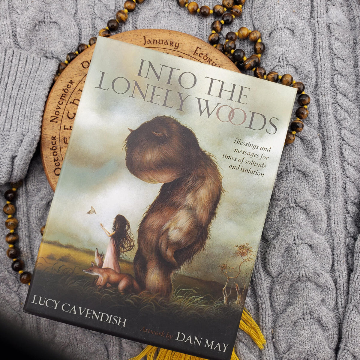 A book titled 'Into the Lonely Woods' by Lucy Cavendish, arranged on a gray cable-knit sweater with a wooden calendar slice and a beaded necklace as props.