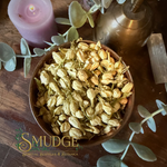 A wooden bowl filled with dried jasmine flowers, styled on a dark wooden surface with a purple candle, eucalyptus leaves, a brass vessel, and a white salt burner. The brand logo 'SMUDGE' is visible in the bottom-left corner.