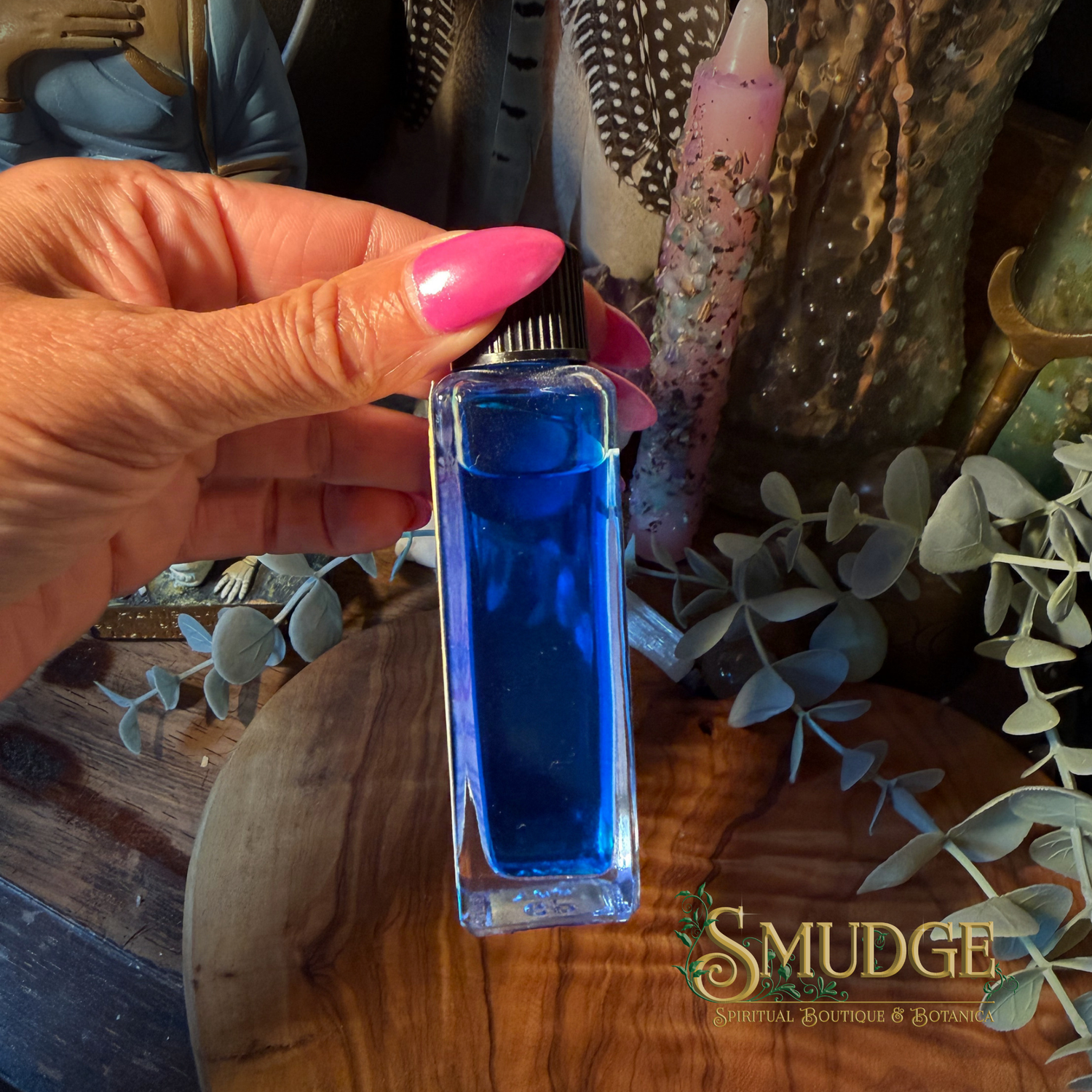 A hand with bright pink nails holds a small rectangular glass bottle filled with vivid blue liquid. The bottle has a black cap and is photographed over a wooden surface with decorative dried leaves and candles in the background.
