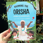 A hand holds a children's book titled 'Knowing the Orisha Gods & Goddesses' against a dark background with blurred greenery. The cover shows an illustrated woman with raised arms, two children, and a bright blue sky background.