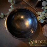 A dark brown wooden bowl with a polished finish, photographed from a top-down angle on a dark wooden surface decorated with eucalyptus leaves. The brand logo 'SMUDGE' is visible in the bottom right corner.