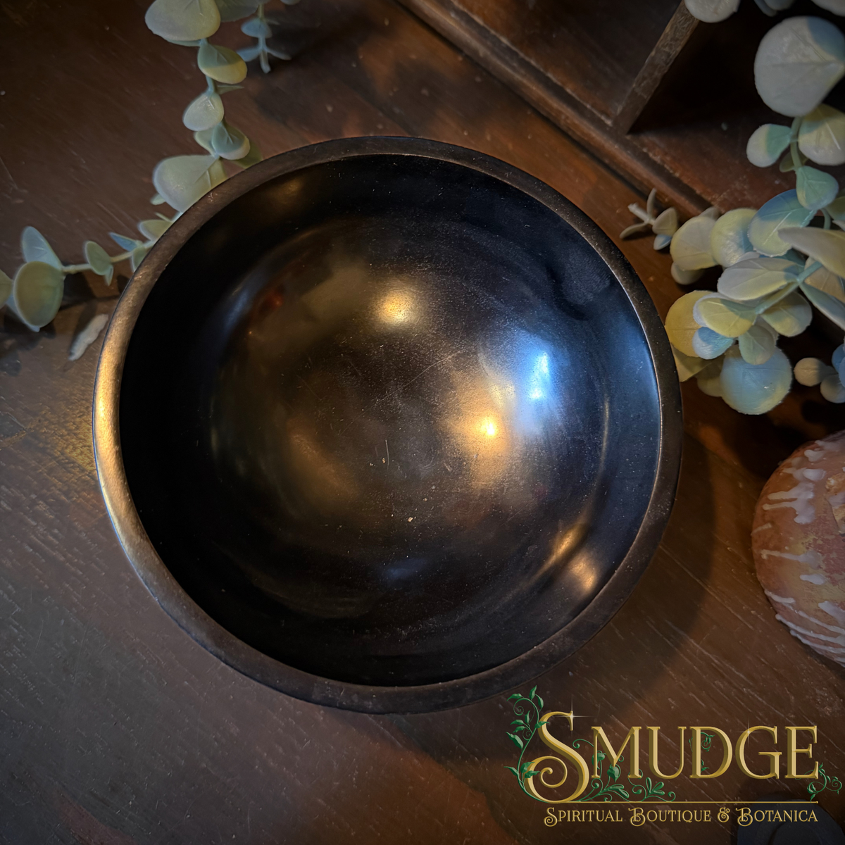 A dark brown wooden bowl with a polished finish, photographed from a top-down angle on a dark wooden surface decorated with eucalyptus leaves. The brand logo 'SMUDGE' is visible in the bottom right corner.