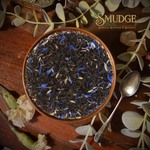 A top-down view of a brown ceramic bowl filled with dark loose-leaf tea mixed with blue flower petals, set on a wooden surface with a spoon, dried flowers, and greenery. The brand logo 'The S Mudge' is visible in the top right.