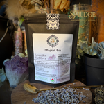 A black stand-up pouch of SMUDGE Magical Tea Lavender Earl Grey is displayed on a wooden surface with loose lavender buds and a spoon. The background features decorative plants, crystals, and a glass jar, with the SMUDGE brand logo visible in the top right corner.
