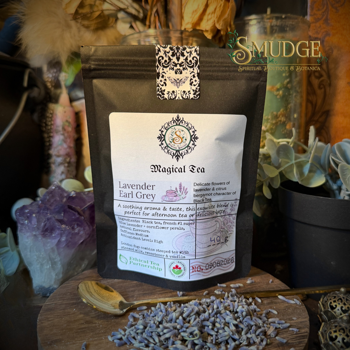 A black stand-up pouch of SMUDGE Magical Tea Lavender Earl Grey is displayed on a wooden surface with loose lavender buds and a spoon. The background features decorative plants, crystals, and a glass jar, with the SMUDGE brand logo visible in the top right corner.