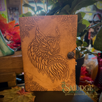 A tan leather journal featuring an embossed owl design on the cover, secured with a dark brown button and cord closure, displayed in a styled setting with plants and autumn leaves.
