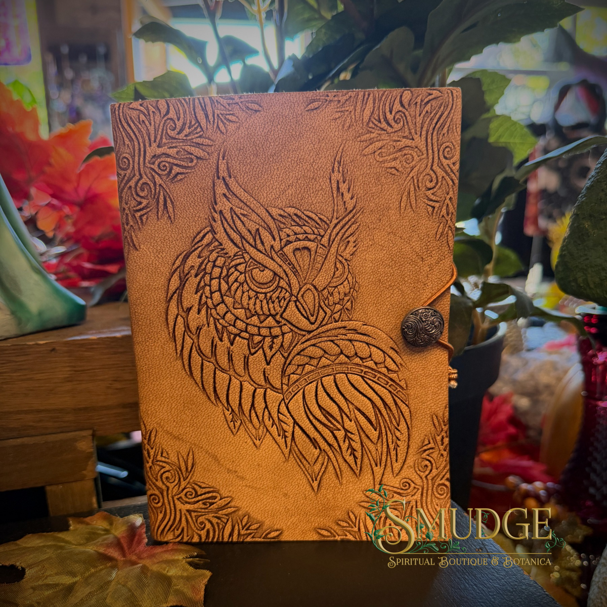 A tan leather journal featuring an embossed owl design on the cover, secured with a dark brown button and cord closure, displayed in a styled setting with plants and autumn leaves.
