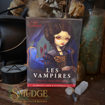A boxed card deck titled "Les Vampires" by Lucy Cavendish stands on a wooden surface surrounded by occult-themed props, including a black cauldron, crystals, dried flowers, and a green glass jar. The box art shows a dark-haired woman in ornate clothing with red eyes.