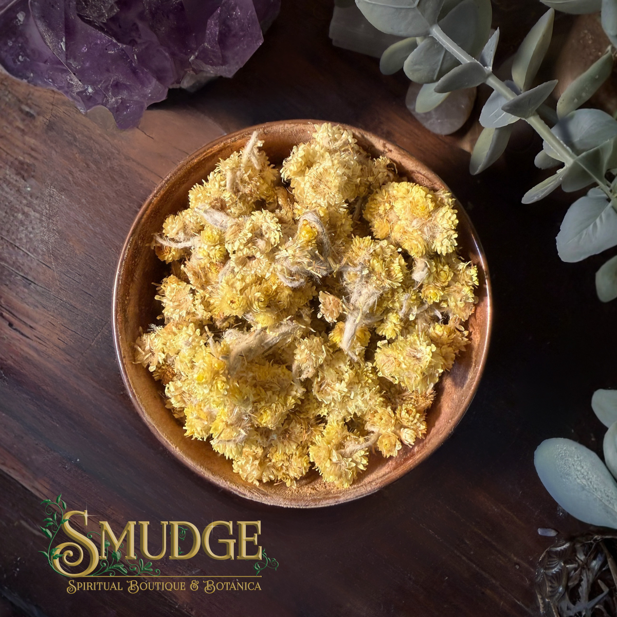 A top-down view of dried yellow flowers, likely chamomile, piled in a small brown wooden bowl. The bowl is set on a dark wooden surface with decorative elements including a purple crystal, eucalyptus leaves, and a logo for 'Smudge Spiritual Boutique & Botánica' in the bottom-left corner.