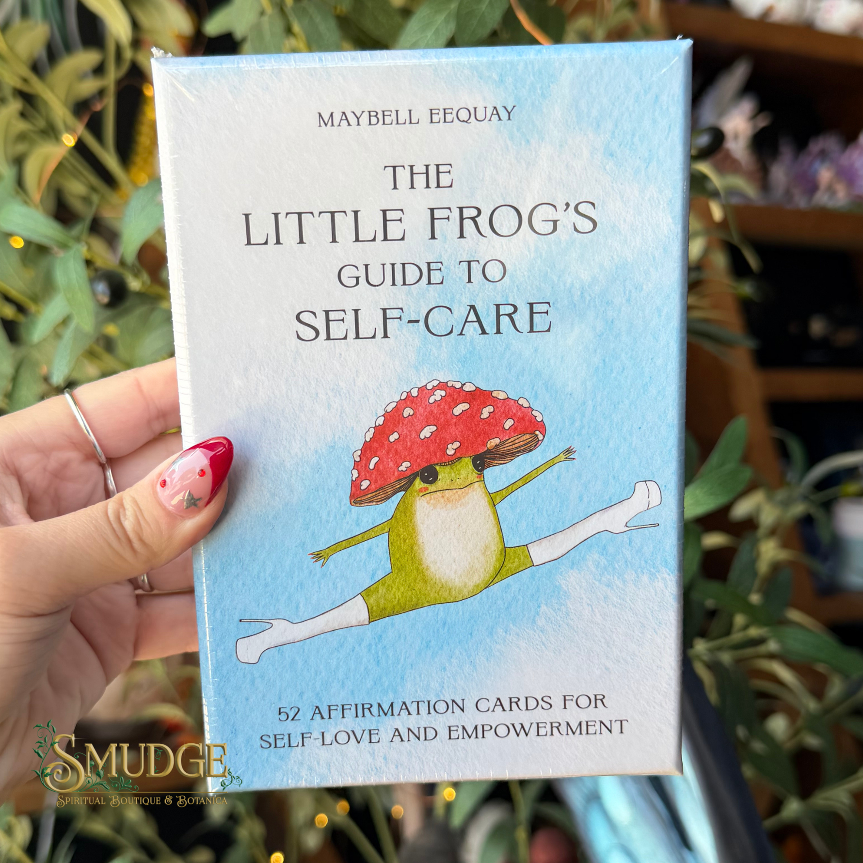 A hand holds a boxed card deck titled "The Little Frog's Guide to Self-Care" by Maybell Eeqouy. The box is light blue with a watercolor-style illustration of a frog wearing a red-and-white mushroom cap and white socks.