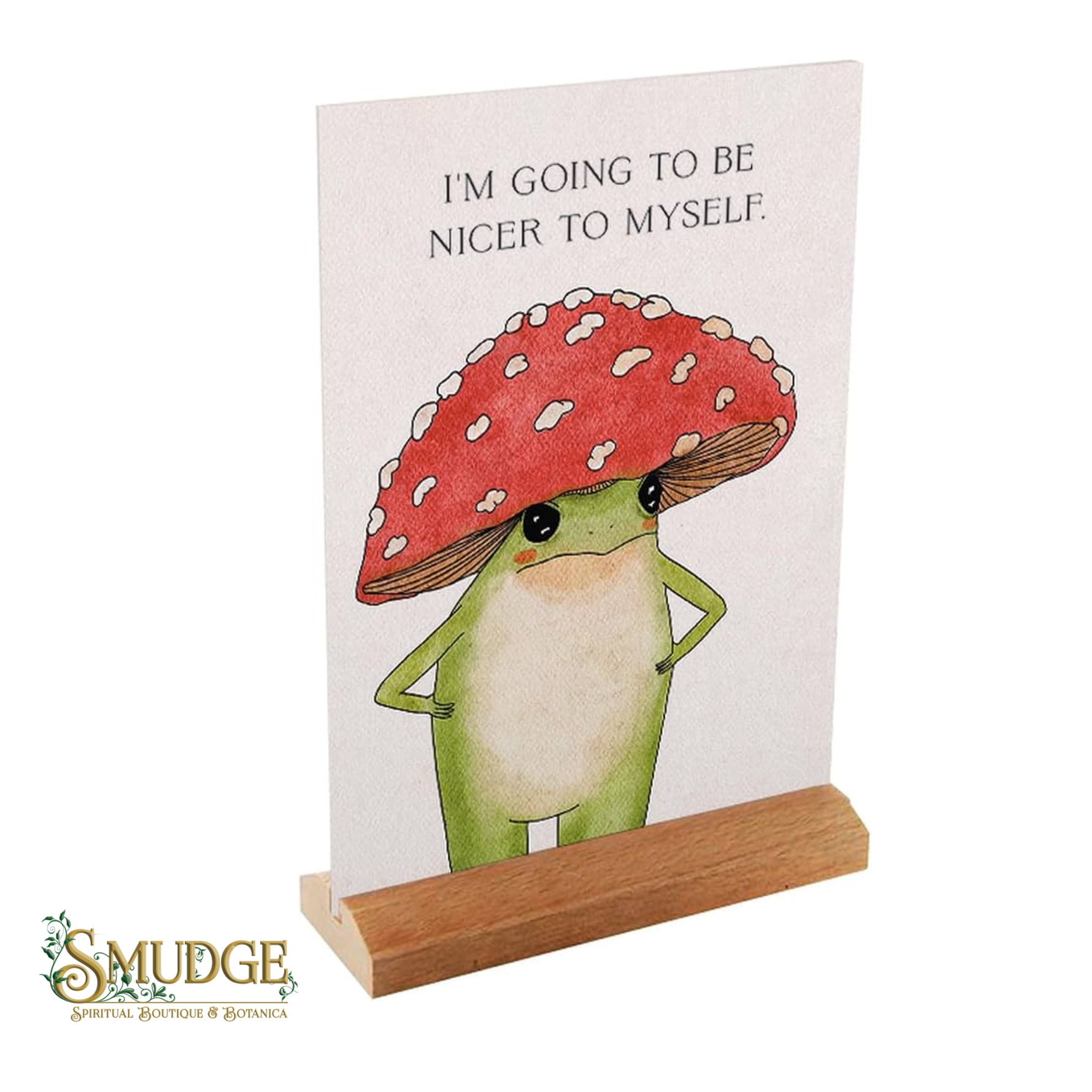 A vertical printed card or sign on a small wooden stand, featuring a cartoon green frog under a red-and-white mushroom with the text 'I'M GOING TO BE NICER TO MYSELF.' A 'SMUDGE Spiritual Boutique & Botanica' logo appears in the lower left corner.
