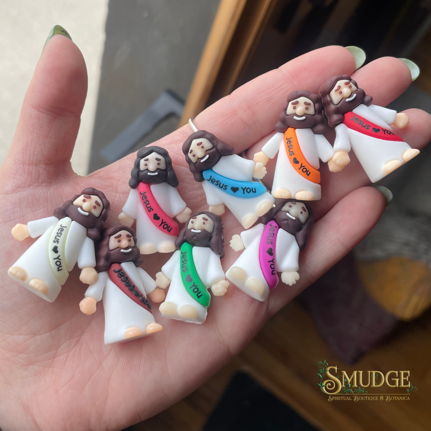 Hand holding small figurines with 'Jesus loves you' messages, brand 'SMUDGE' visible.