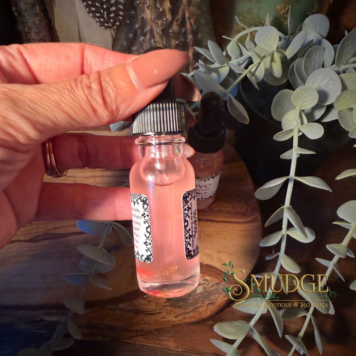 A hand holds a small clear glass dropper bottle containing pink liquid, love & attraction oil, with a black-and-white patterned label and a black dropper cap. Another similar bottle sits in the background on a wooden surface, surrounded by green foliage.