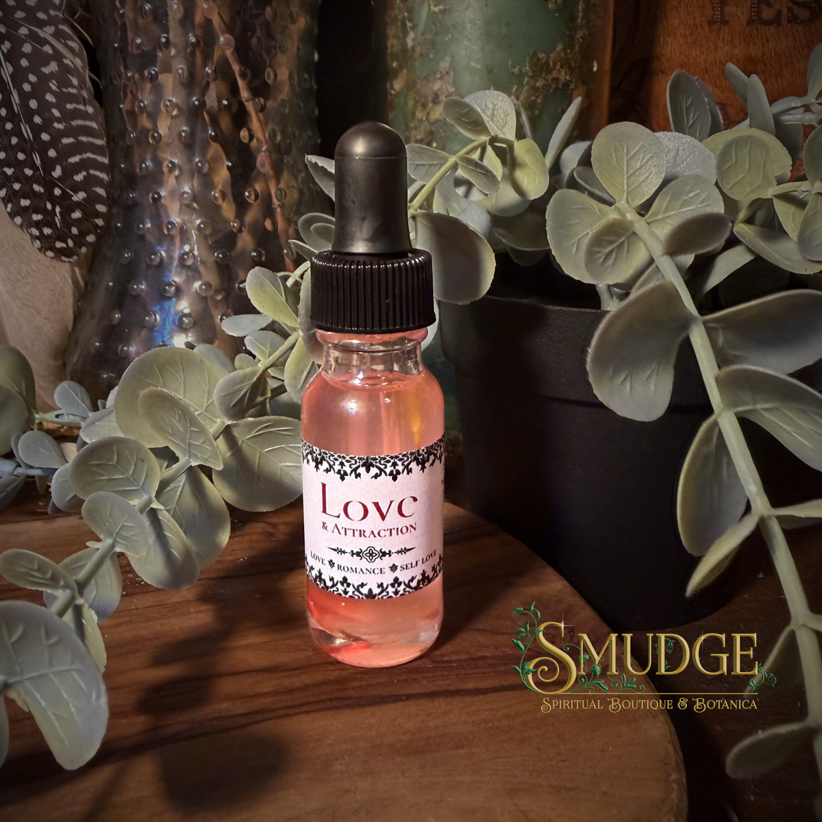 A small glass dropper bottle filled with pink liquid sits on a wooden surface surrounded by green leaves and dried botanical decor. The label reads 'Love & Attraction Oil' and the bottom-right corner shows the brand name 'SMUDGE'.