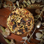 A top-down view of a rustic brown bowl filled with a colorful herbal tea blend, surrounded by dried flowers, green leaves, and a silver spoon on a dark wooden table. The brand logo 'SMUDGE BOTANICA' is visible in the top right corner.