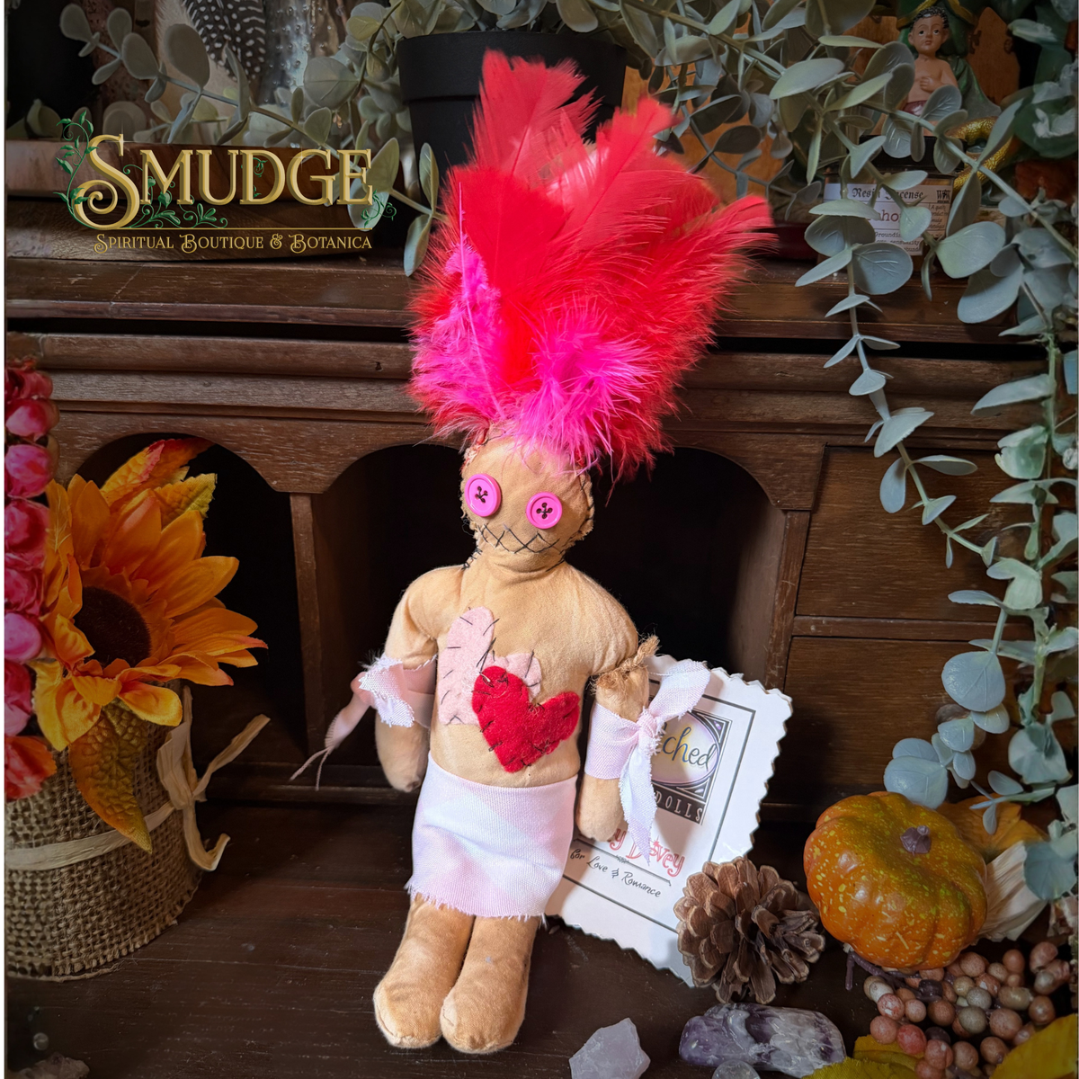A handmade wooden doll with a bright pink feather headdress, pink bow details, and a red heart patch on its chest, styled on a wooden mantel with autumn decorations and a 'Smudge Society' logo in the background.