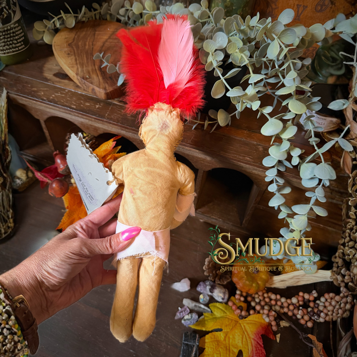 A handmade cloth doll with tan fabric, a red feather headdress, and a white waistband is being held by a hand. The background features a rustic wooden shelf decorated with eucalyptus, autumn leaves, and dried botanicals.
