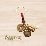 A brass keychain featuring a red braided cord, a gold split ring, a Chinese coin charm, a lucky cat charm, a gourd charm, and a engraved brass tag, displayed on a light wood background with the 'SMUDGE' brand logo in the lower-left corner.